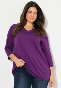 Lila V-Ausschnitt-Oberteil mit Dreiviertelärmeln, lockerer Schnitt und leicht schrägem Saum. Glatte Textur mit leichtem Glanz.
