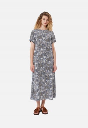 Donna con i capelli ricci che indossa un abito midi blu a fiori con maniche corte e sandali marroni con frange, in piedi su sfondo bianco.