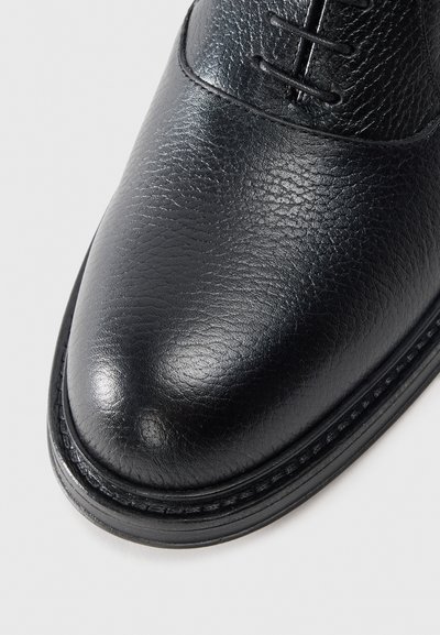 Black leather shoe with a textured finish, rounded toe, and contrasting stitching along the sole. Laces are present on the upper.
