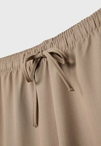 Beige elastische tailleband met een strik aan de voorkant, gemaakt van een gladde stof. De textuur lijkt zacht en lichtgewicht.