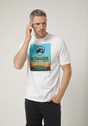 Hombre con cabello corto que lleva una camiseta blanca con una montaña colorida y texto "ALTONADOCK FARO DE LARIÑO", tocándose la oreja con una mano.