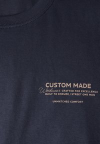 Marinblå t-shirt med rund hals, försedd med ljusbeige text som säger "CUSTOM MADE" och andra detaljer, i mjukt bomullsmaterial.