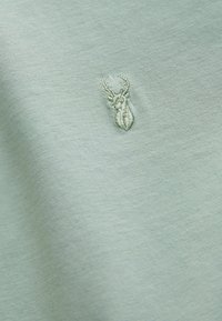 Világoszöld textil sima felülettel, melyen hímzett szarvas fej design található, azonos színű cérnával. Nagyított nézet.