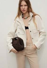 Crème denim jack met twee voorzakken, gecombineerd met beige nauwsluitende broek. Bruin gevlochten tas in de hand, met een ronde vorm.