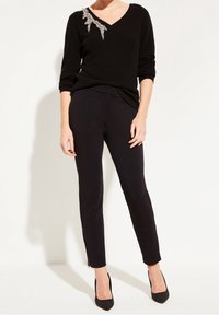 Un pull noir côtelé avec un col en V et des embellissements en argent sur l'épaule, associé à un pantalon slim noir et des talons texturés noirs.