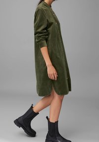 Persona che indossa un vestito a camicia in velluto a coste verde oliva con maniche lunghe arrotolate e stivali neri alla caviglia, che cammina su uno sfondo grigio.