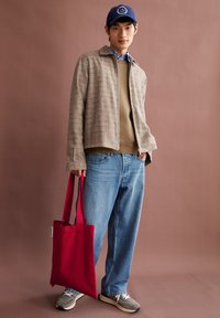 Jonge man met een blauwe pet, beige gestructureerde jas, bruine trui, blauwe jeans, grijze sneakers, die een rode draagtas vasthoudt tegen een bruine achtergrond.