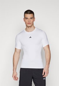 Hvid kortærmet træningsshirt lavet af strækbart materiale, med rund halsudskæring og et lille sort Adidas-logo på brystet.