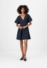 Vestido azul marino con escote en V, mangas cortas, falda acampanada y un corsé ajustado. Lleva unos tacones negros con pulsera en el tobillo. Textura suave.