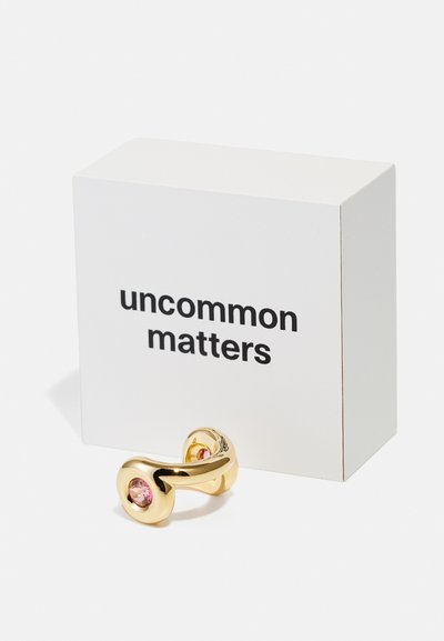 Anello color oro con una forma liscia e curva, decorato con due gemme rosa incastonate; confezionato in una scatola bianca con la scritta "uncommon matters".
