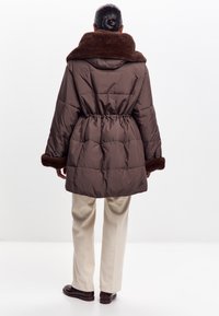 Abrigo acolchado marrón con un amplio cuello de piel sintética, cinturilla elástica y puños de piel marrón en contraste. Usado sobre pantalones de color claro.