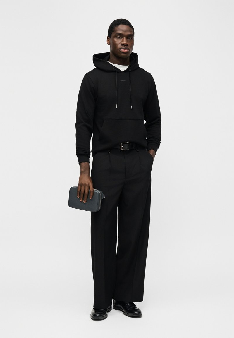 Mann steht, trägt schwarzen Kapuzenpullover, schwarze weit geschnittene Hose, schwarzen Gürtel, schwarze Schuhe, hält eine dunkelgraue Clutch in der linken Hand vor einfarbigem Hintergrund.