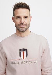Felpa rosa chiaro con logo a scudo con "1924" e strisce verticali rosse, bianche e blu. Design semplice con colletto a girocollo. Tessuto morbido.