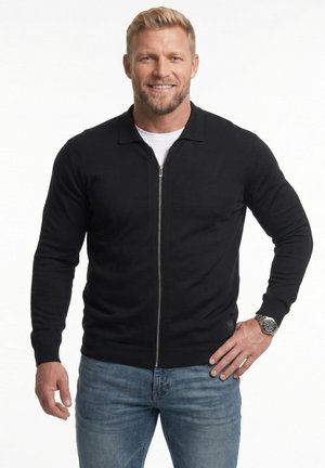 Blonder Mann lächelt, trägt einen schwarzen Reißverschluss-Pullover, weißes Unterhemd, blaue Jeans und eine silberne Armbanduhr, steht mit einer Hand an der Hüfte.