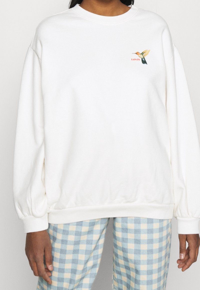 Femme portant un sweat-shirt blanc avec un petit colibri coloré et le logo rouge "LEVI'S" sur la poitrine, associé à un pantalon à carreaux bleu et blanc.