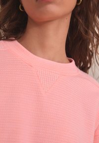 Light pink textured sweatshirt with a round neck and triangular accent at the shoulder. Fabric appears soft, featuring a subtle pattern.