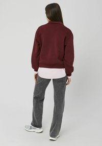 Sweat-shirt bordeaux avec col montant, ourlet à côtes, superposé à une chemise rayée rose clair, associé à un pantalon gris et des chaussures de sport.