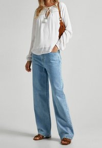 Donna che indossa una blouse bianca a maniche lunghe, jeans a gamba larga blu chiaro, sandali marroni e borsa a tracolla marrone, in piedi su uno sfondo semplice.