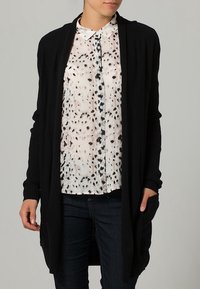 Donna che indossa un cardigan nero aperto davanti su una camicia bianca con macchie nere e jeans blu scuro, mani rilassate ai lati.