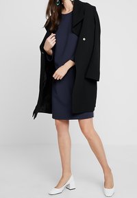 Cappotto oversized nero, vestito blu navy e décolleté bianchi con tacco a blocco. Maniche lunghe, scollatura rotonda, tessuto morbido con vestibilità ampia.