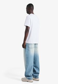 Bela majica s kratkimi rokavi, združena s svetlo modrimi, rahlo ohlapnimi kavbojkami z gladkim prehodom barve od temne do svetle in nevtralnimi supergami.