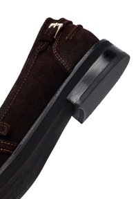 Scarpa in suede marrone con cuciture a contrasto, dettaglio con fibbia laterale e suola in gomma nera. Tacco leggermente elevato e forma arrotondata.