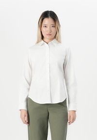 Woman with straight long hair wearing a white buttoned long-sleeve shirt and olive green pants, standing against a plain white background.