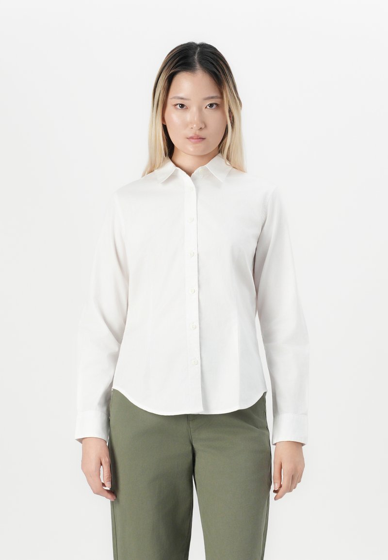 Woman with straight long hair wearing a white buttoned long-sleeve shirt and olive green pants, standing against a plain white background.