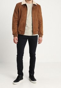 Giacca in suede marrone con colletto in ecopelliccia beige, pantaloni neri slim-fit e sneakers nere con suola bianca, in piedi su un pavimento semplice.