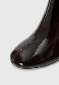 Botín de tobillo burdeos brillante con punta afilada, textura suave, costuras decorativas y un diseño ajustado, mostrando una elegancia estilizada.