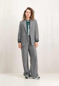 Grijze op maat gemaakte pak met wijde pijpen en een bijpassende blazer. Daaronder een groene trui die over een gestreept shirt is gedragen. Opvallende sneakers.