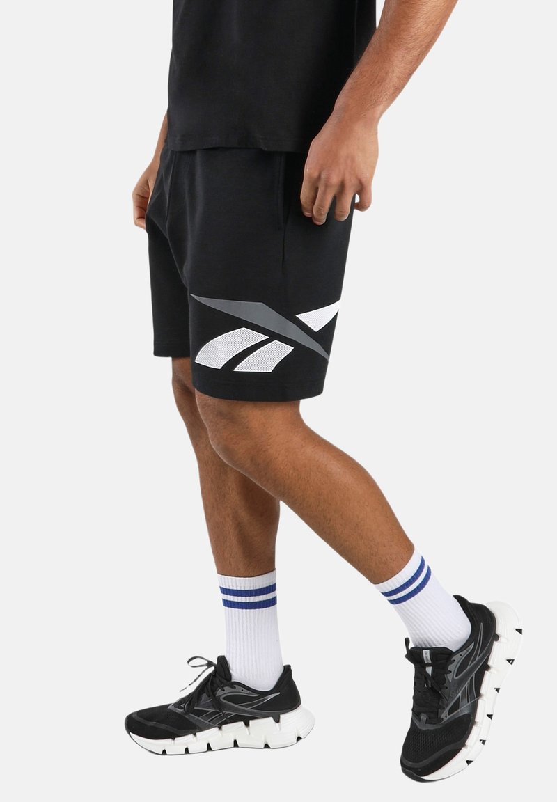 Man wearing black athletic shorts with white and gray logo, white socks with blue stripes, and black running shoes, standing on one foot.