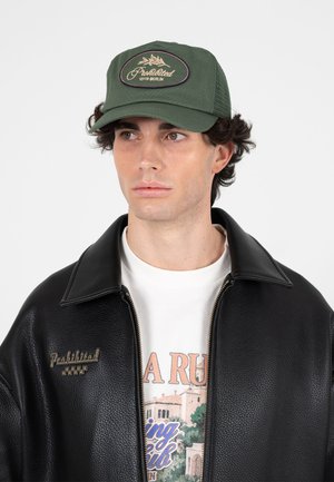 Jonge man met een groene pet met de tekst "Prohibited 10119 Berlin" en een zwarte leren jas over een wit shirt met print, kijkt opzij.