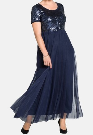 Woman wearing a navy blue gown with a sequined bodice and sheer flowing skirt, lifting the hem slightly with one hand.