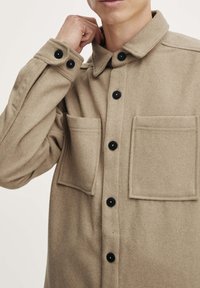 Beige Wollhemdjacke mit Kragen, schwarzen Knöpfen und zwei Fronttaschen. Der Stoff hat ein weiches, strukturiertes Aussehen.