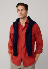 Rote Knopfleiste-Hemd aus strukturiertem Material mit einem dunkelblauen Strickpullover, der über die Schultern drapiert ist; hellgraue karierte Hose.