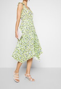 Frau in einem gelben, blumigen, plissierten Midikleid mit gebundenen Schulterträgern, weißen Riemchensandalen und einer weißen Clutch in der Hand.