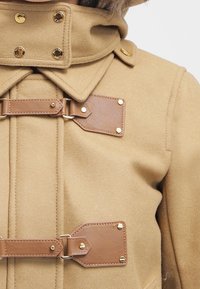 Close-up van een camel-kleurige jas met leren togglesluiting en gouden drukknoopjes op de kraag en de voorclosure.