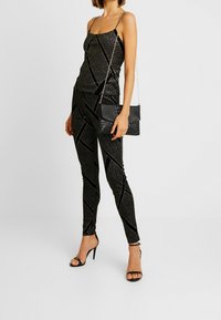 Camisola nera lucida e leggings a vita alta abbinati con linee diagonali testurizzate. Abbinati a una mini borsa nera con texture e tacchi alti.
