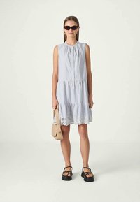 Kvinne i en ermeløs lys blåstripete kjole med blonder nederst, svarte plattformsandaler, brun håndveske og mørke solbriller, som står foran en hvit bakgrunn.