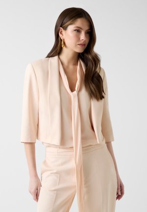 Vrouw met lang bruin haar die een beige cropped blazer, bijpassende broek en blouse met strikdetail aan de voorkant draagt, staand tegen een witte achtergrond.