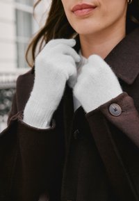 Guanti bianchi lavorati a maglia realizzati in materiale morbido, con una vestibilità aderente e polsini a coste, indossati da una persona con un cappotto marrone scuro.