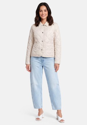 Vrouw draagt een beige gewatteerde jas, lichtblauwe spijkerbroek en witte open-toe hakken, staand en glimlachend tegen een effen achtergrond.