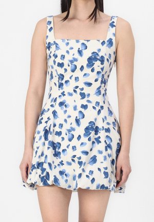Femme portant une robe blanche sans manches avec des motifs floraux bleus et un décolleté carré, debout devant un fond blanc uni.