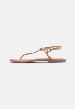 Sandalias planas doradas con tiras trenzadas, hebilla ajustable en el tobillo y suela beige. Diseño simple con punta abierta y detalles mínimos.