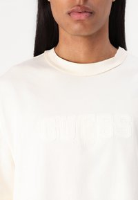 Kremfarget bomulls T-skjorte med "GUESS ORIGINALS" preget tekst på forsiden, rund hals, og kortermet med en lett tekstur.