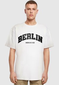 Witte katoenen t-shirt met korte mouwen, met "BERLIN" in vetgedrukte zwarte letters en "MERCH•DE" eronder in kleinere letters.