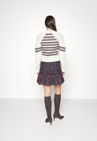 Femme aux longs cheveux noirs portant un pull blanc à motifs, une jupe fleurie bleu marine et des bottes noires montantes à talons, debout face à un mur blanc.