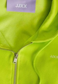Neonově zelená mikina na zip vyrobená z měkké tkaniny, se stříbrným zipem a fialovou etiketou s textem "JJXX". Hladký povrch a přiléhavý střih.