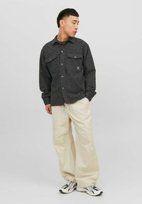 Giacca a camicia grigia a scacchi con due tasche sul petto, chiusura con bottoni e vestibilità rilassata, abbinata a pantaloni beige larghi e sneakers.